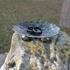 Pode incluir: Um porta-anéis de metal preto com acabamento martelado. O suporte tem um pequeno prato e três pernas. Duas alianças pretas estão descansando no prato.