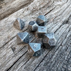 Könnte beinhalten: Ein Set aus sieben polyedrischen Würfeln aus dunklem, verwittertem Metall mit abgenutzter Oberfläche. Die Sammlung umfasst verschiedene Formen wie vier-, sechs-, acht-, zehn-, zwölf- und zwanzigseitige Würfel, die alle mit schwarzen Zahlen versehen sind.