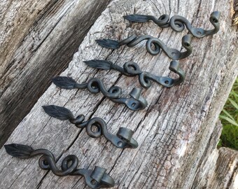 May include: A set of five dark gray metal wall hooks, each featuring a forged leaf-shaped end, a central spiral design, and a curved hook at the opposite end. Each piece includes a small circular hole for mounting, showcasing a textured finish.