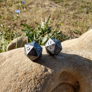 May include: Two silver metal dice with black numbers on each side. The dice are sitting on a light brown rock.