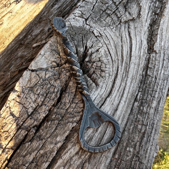A real blacksmith hammered this bottle opener from iron — the hand-forged texture and heft make it feel like a relic from a 19th-century forge, not a gas station gift shop.