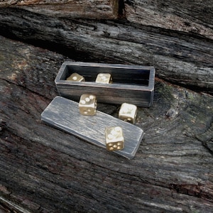 Puede incluir: Un juego de dados de latón con una caja y una bandeja de madera. Los dados son de color dorado con puntos negros. La caja y la bandeja son de madera gris oscuro. Los dados están colocados en la bandeja y en la caja.