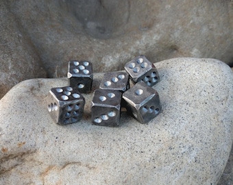 Può includere: Un insieme di sei piccoli dadi in metallo grigio scuro con una texture ruvida e martellata. Ogni cubo presenta dei punti circolari incavati che rappresentano i numeri, con variazioni visibili nel posizionamento e nella profondità dei segni su ogni dado.