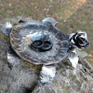 Peut inclure: Un porte-alliance en métal en forme de tortue avec une rose sur le côté. La tortue a un dos en forme de plat pour tenir les alliances. Les alliances sont noires et sont posées dans le plat.