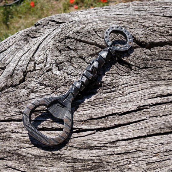 A blacksmith literally hammers this bottle opener out of raw metal — you can see the forge marks, no two are identical.