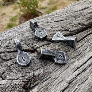 Puede incluir: Cuatro piezas de juego individuales de metal con un acabado oscuro y desgastado. Cada pieza presenta una forma geométrica distinta, incluyendo un círculo, un hexágono, un triángulo y un rectángulo, con números en relieve en la superficie.