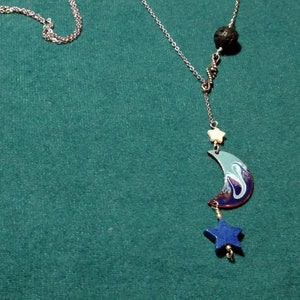 May include: A silver chain necklace with a blue and green crescent moon charm, a white star charm, and a blue star charm. The necklace also features a black lava rock bead.