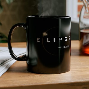 May include: Black ceramic mug with the word "ECLIPSE" and the date "08.12.2026" printed in white. The mug is on a wooden surface, with steam rising from it. A glass teapot is in the background.