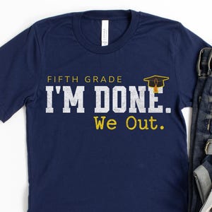 May include: A navy blue t-shirt with the text "FIFTH GRADE I'M DONE. We Out." in white and yellow. The shirt is styled with blue denim jeans and white trainers, creating a casual outfit. A graduation cap graphic is included.