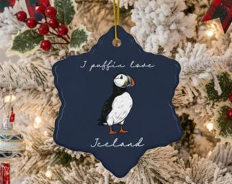 May include: A Christmas tree decorated with ornaments, including a navy blue, star-shaped ornament featuring a puffin illustration and the text "I puffin love Iceland." The tree also has red and black plaid bows, red berries, and blue and white ornaments.
