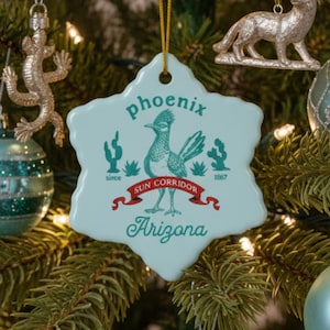 Peut inclure: Un sapin de Noël décoré avec des boules de Noël teal et argentées, ainsi que des ornements décoratifs en forme de lézard, de cerf et de cactus. Un ornement bleu clair avec le texte "Phoenix Arizona" est au centre.