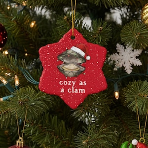 May include: A decorated Christmas tree with red and gold ornaments, white snowflakes, and string lights. A red, star-shaped ornament with a clam design and the text "cozy as a clam" hangs from the tree.