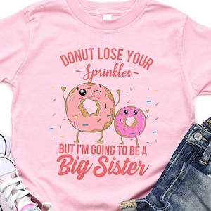 Puede incluir: Una camiseta rosa para niños pequeños con el texto "Donut Lose Your Sprinkles" y "But I'm Going to Be a Big Sister" con gráficos de donuts. La imagen también incluye un par de zapatillas rosas y vaqueros azules doblados.
