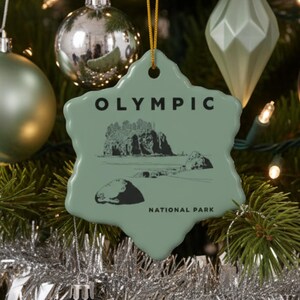 May include: A decorated Christmas tree with various ornaments. The ornaments include silver, matte green, and dark gray spheres, diamond shapes, and a star-shaped ornament with "OLYMPIC NATIONAL PARK" printed on it. Silver tinsel and lights are also visible.
