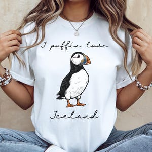 May include: A white t-shirt featuring a graphic illustration of a puffin with black, white, and orange coloring. The text "I puffin love" is printed in black cursive above the bird, and the word "Iceland" is printed in black cursive below it.