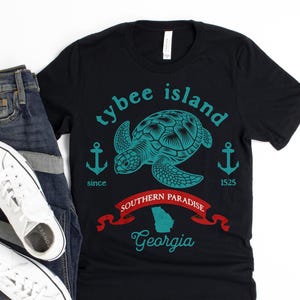 May include: A black short-sleeve t-shirt featuring a teal sea turtle graphic, two anchors, and the text Tybee Island since 1525. Below is a red banner with the text Southern Paradise, a teal outline of Georgia, and the word Georgia in teal script.