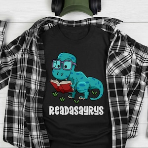 Puede incluir: Camiseta negra con un dinosaurio azul que lleva gafas y lee un libro rojo. El texto "READASAURUS" está impreso debajo del dinosaurio.