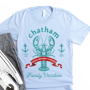 May include: A light blue t-shirt featuring a teal lobster graphic, two anchors, and text that reads Chatham, Cape Cod, est. 1664, Making Memories Together, and Family Vacation. Also included are grey shorts and a pair of white canvas sneakers.