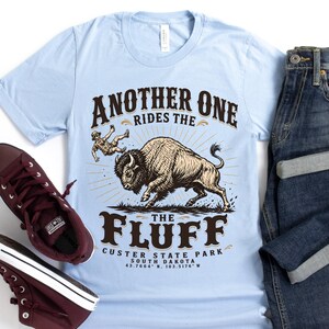May include: A light blue t-shirt featuring a graphic of a bison and a person being tossed, with the text "ANOTHER ONE RIDES THE FLUFF CUSTER STATE PARK SOUTH DAKOTA 43.7864 N, 103.5176 W". Dark blue denim jeans and maroon high-top sneakers are included.