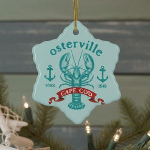 Könnte beinhalten: Ein hellblaues Keramikornament mit gewelltem Rand und einer dunkel-türkisfarbenen Hummer-Illustration in der Mitte. Der Text lautet "osterville" über dem Hummer, "since 1648" an den Seiten und "CAPE COD" auf einem roten Banner darunter.