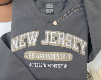 May include: A dark gray crewneck sweatshirt featuring the text NEW JERSEY in a distressed white and tan font, with THE GARDEN STATE and the coordinates 40°13'13"N 74°45'57"W printed below. The garment has long sleeves and a ribbed neckline.