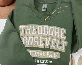 May include: An olive green crewneck sweatshirt featuring the white and tan text Theodore Roosevelt National Park with coordinates 46 N 103 27 W. The garment is displayed alongside a brown felt hat, a gold chain necklace, and a dried palm leaf.