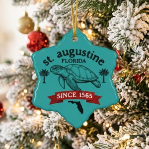 Könnte beinhalten: Ein türkisfarbener Keramikschmuck mit gewelltem Rand, der an einer goldenen Schnur hängt. Er zeigt eine schwarze Illustration einer Meeresschildkröte, Palmen und den Text "st. augustine FLORIDA SINCE 1565" auf einem roten Banner über einer schwarzen Florida-Silhouette.