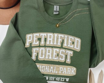 May include: An olive green crewneck sweatshirt featuring the white and tan text PETRIFIED FOREST NATIONAL PARK and coordinates 35°N 109°48'23"W. The garment has long sleeves, a ribbed collar, and a Gildan brand label visible at the inner neckline.