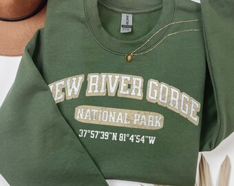 May include: An olive green crewneck sweatshirt featuring the white text NEW RIVER GORGE NATIONAL PARK and the coordinates 37°57'39"N 81°4'54"W printed on the chest. The garment is displayed alongside a brown felt hat and a gold chain necklace.