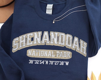 May include: A navy blue crewneck sweatshirt featuring the text SHENANDOAH NATIONAL PARK in a distressed beige font, with the coordinates 38°32'54"N 78°27'38"W printed in white below. The garment has long sleeves and a ribbed collar.