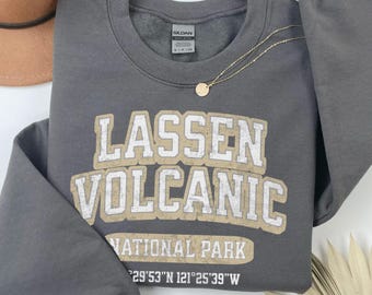 May include: A dark gray long-sleeve sweatshirt featuring the text LASSEN VOLCANIC NATIONAL PARK in a distressed beige and white font, along with the coordinates 40°29'53"N 121°25'39"W printed in white below the main graphic design.