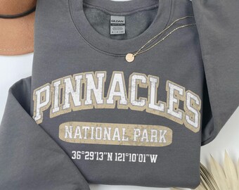 May include: A dark gray crewneck sweatshirt featuring the text PINNACLES NATIONAL PARK in a distressed beige font with a white outline, along with the coordinates 36°29'13"N 121°10'01"W printed in white below the main graphic.