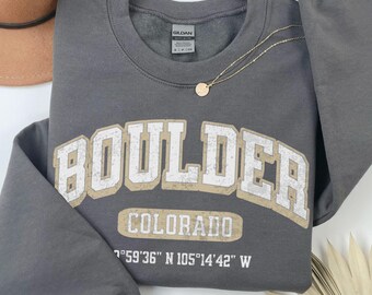 May include: A dark gray long-sleeve crewneck sweatshirt featuring the text BOULDER COLORADO and the coordinates 40°01'07" N 105°16'36" W in white and tan distressed lettering. The garment has a Gildan brand label at the inner neckline.