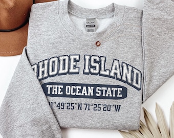 May include: A heather gray crewneck sweatshirt featuring dark blue text that reads RHODE ISLAND THE OCEAN STATE with coordinates 41°49'25"N 71°25'20"W printed below. The garment has long sleeves and a Gildan brand label at the inner neckline.
