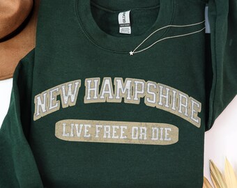 May include: A dark forest green crewneck sweatshirt featuring the text NEW HAMPSHIRE in a curved, distressed light tan font, with the state motto LIVE FREE OR DIE printed in a matching rectangular block below the main lettering.