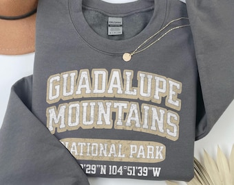 May include: A dark gray crewneck sweatshirt featuring the white and tan text Guadalupe Mountains National Park and the coordinates 29°N 104°51'39"W. The garment has long sleeves and a ribbed collar, with a Gildan brand tag visible at the neckline.