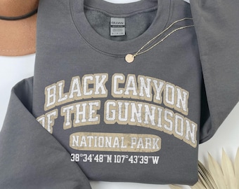 May include: A charcoal gray crewneck sweatshirt featuring the text Black Canyon of the Gunnison National Park in a distressed beige and white font, along with the coordinates 38 34 48 N 107 43 39 W printed in white on the front.
