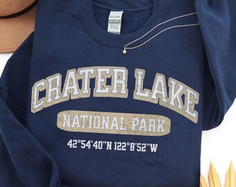 May include: A navy blue crewneck sweatshirt featuring the text CRATER LAKE NATIONAL PARK in a distressed beige font, along with the coordinates 42°54'40"N 122°8'52"W printed in white below the text. The garment has long sleeves and a ribbed collar.