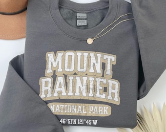 May include: A dark gray long-sleeve crewneck sweatshirt featuring the white and tan text MOUNT RAINIER NATIONAL PARK and the coordinates 46°51'N 121°45'W. The garment has a Gildan brand tag at the inner neckline and a gold-toned chain necklace.