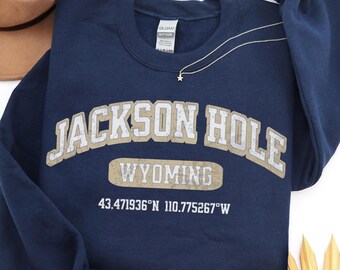 May include: A navy blue crewneck sweatshirt featuring the text JACKSON HOLE WYOMING in a distressed beige font, along with the coordinates 43.471936°N 110.775267°W printed in white below the text. The garment has long sleeves and a ribbed collar.