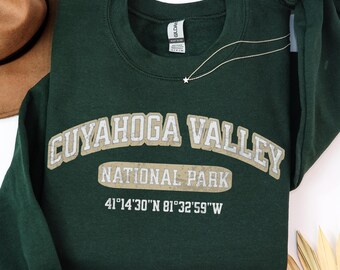 May include: A dark green crewneck sweatshirt featuring the text CUYAHOGA VALLEY NATIONAL PARK in a beige and white collegiate font, with the coordinates 41°14'30"N 81°32'59"W printed below in white. The garment has long sleeves and a ribbed collar.
