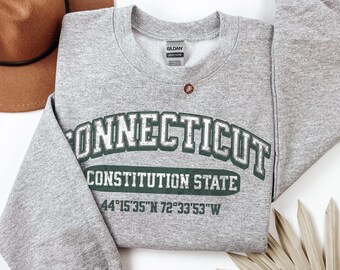 May include: A heather gray crewneck sweatshirt featuring dark green text that reads CONNECTICUT CONSTITUTION STATE and the coordinates 44°15'35"N 72°33'53"W. The garment has long sleeves, a ribbed neckline, and a Gildan brand label at the collar.