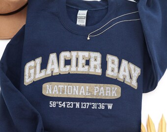 May include: A navy blue crewneck sweatshirt featuring the text GLACIER BAY NATIONAL PARK in a distressed beige font, along with the coordinates 58°54'23"N 137°31'36"W printed in white below the text. The garment has long sleeves and a ribbed collar.