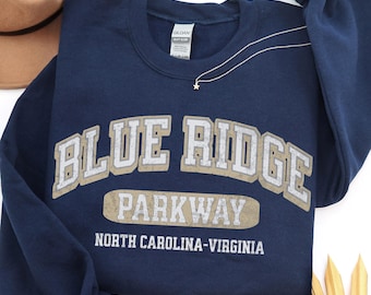 May include: A navy blue crewneck sweatshirt featuring the text BLUE RIDGE PARKWAY NORTH CAROLINA-VIRGINIA in a distressed beige and white font. The garment has long sleeves and a ribbed collar, with a Gildan brand label visible at the inner neckline.