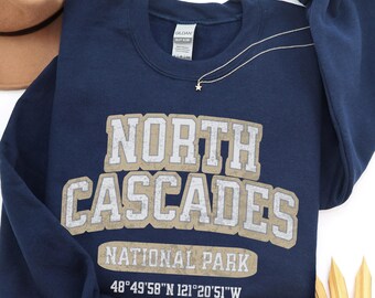 May include: A navy blue long-sleeve sweatshirt featuring the text NORTH CASCADES NATIONAL PARK and coordinates 48°49'58"N 121°20'51"W in a distressed white and tan font. The garment has a crew neckline and a Gildan brand label at the collar.