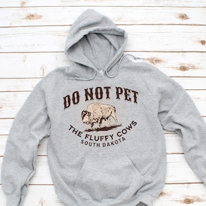 May include: Grey hooded sweatshirt with a brown graphic of a bison and the text "Do Not Pet The Fluffy Cows South Dakota".