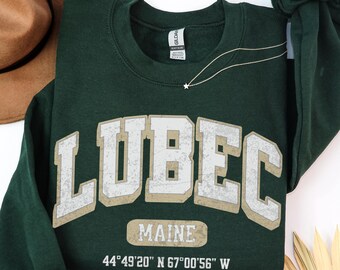 May include: A dark green long-sleeve crewneck sweatshirt featuring the text LUBEC in large white block letters with a distressed finish. Below, the word MAINE and the coordinates 44°49'20" N 67°00'56" W are printed in white.