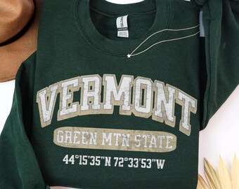 May include: A dark forest green crewneck sweatshirt featuring the text VERMONT in large arched letters, with GREEN MTN STATE and the coordinates 44°15'35"N 72°33'53"W printed below in a distressed, light-colored, textured font style.