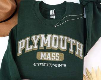 May include: A dark green crewneck sweatshirt featuring the text PLYMOUTH MASS in a distressed white and gold font, with the coordinates 41°57'30"N 70°40'04"W printed below in white. The garment has long sleeves and a ribbed collar.