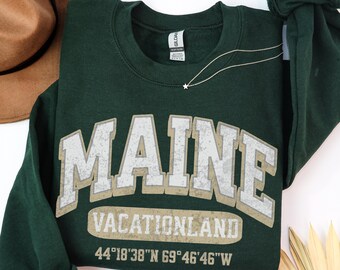 May include: A dark green crewneck sweatshirt featuring the word MAINE in large, distressed, light-colored block letters. Below, the text VACATIONLAND appears in a smaller font, followed by the coordinates 44°18'38"N 69°46'46"W in white.